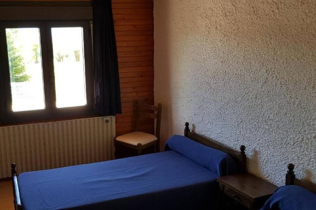 A bedroom with a wooden floor, a bed with a blue bedspread, a chair, a small table, and a window with curtains.