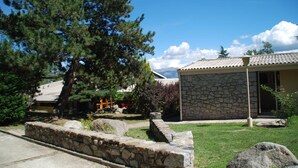 Un bâtiment en pierre, surmonté d’un toit en tuiles, entouré d’un mur de pierre et d’une zone herbeuse plantée d’arbres.