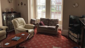 A living room with a sofa, armchair, and a coffee table. There is a TV on a stand and a bookshelf. - Viking Guest House (Bridlington)