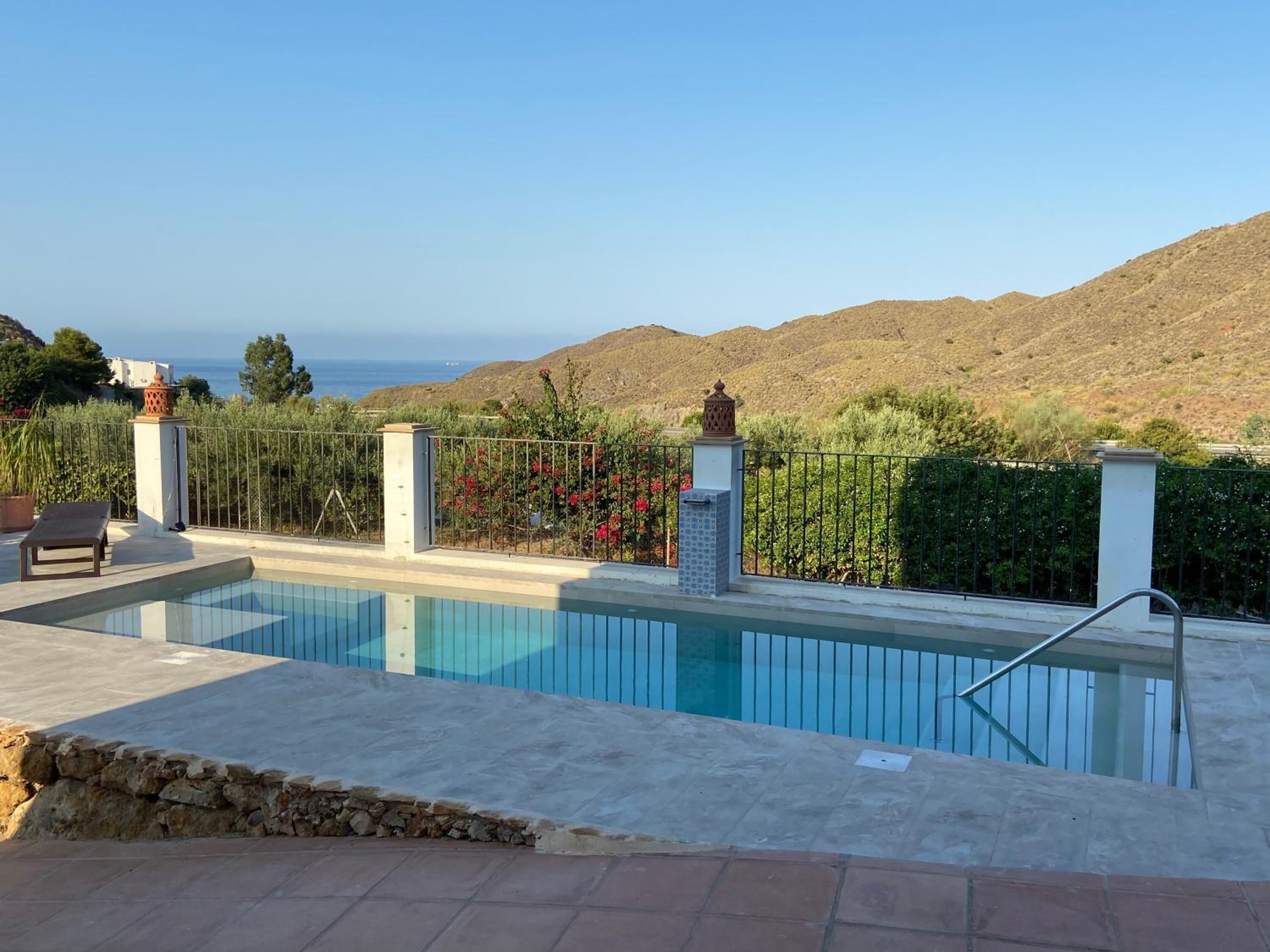 Une piscine bordée de pierres, entourée d’une clôture métallique, et offrant une vue sur la mer et les collines.
