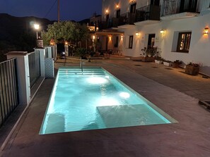 Une piscine extérieure à l’eau claire, entourée d’une zone dallée agrémentée de plantes en pot et d’un bâtiment doté de nombreux balcons et d’un éclairage extérieur.