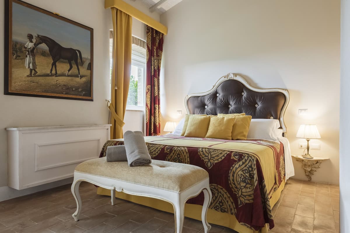 Une chambre à coucher avec un grand lit, un banc, un tableau et une fenêtre avec des rideaux.