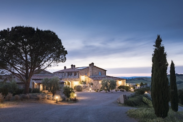 A stone house with a gravel driveway, surrounded by trees and shrubs, at dusk.