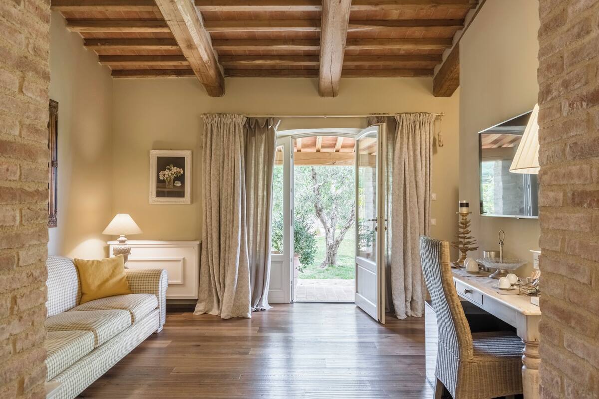 Une pièce chaleureuse avec un plafond en bois, un canapé agrémenté d’un coussin, un bureau avec une chaise, et une vue sur un jardin par une porte ouverte.