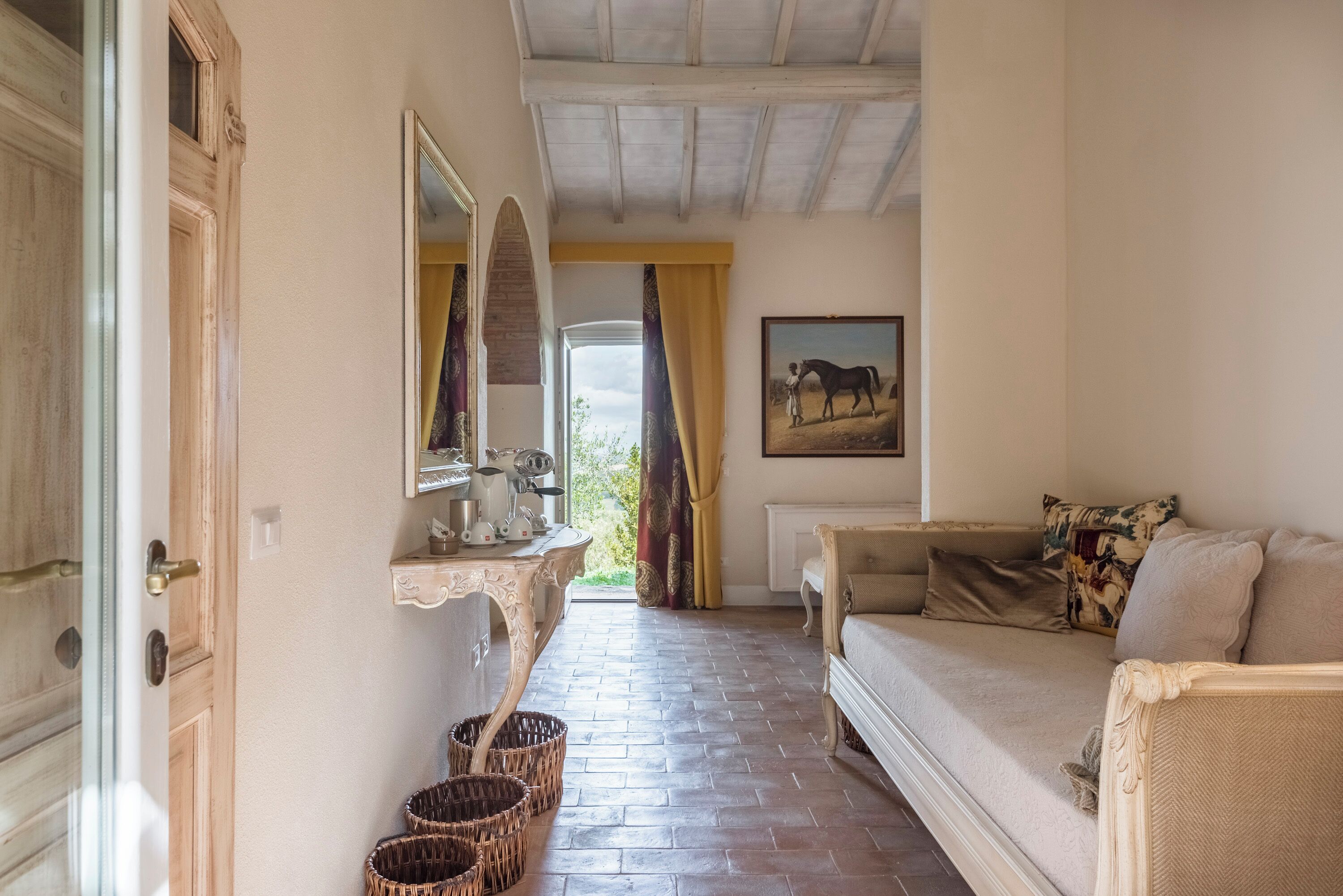 A hallway with a wooden door, a mirror, a table with a pitcher and cups, wicker baskets, a sofa, and a painting of a horse.
