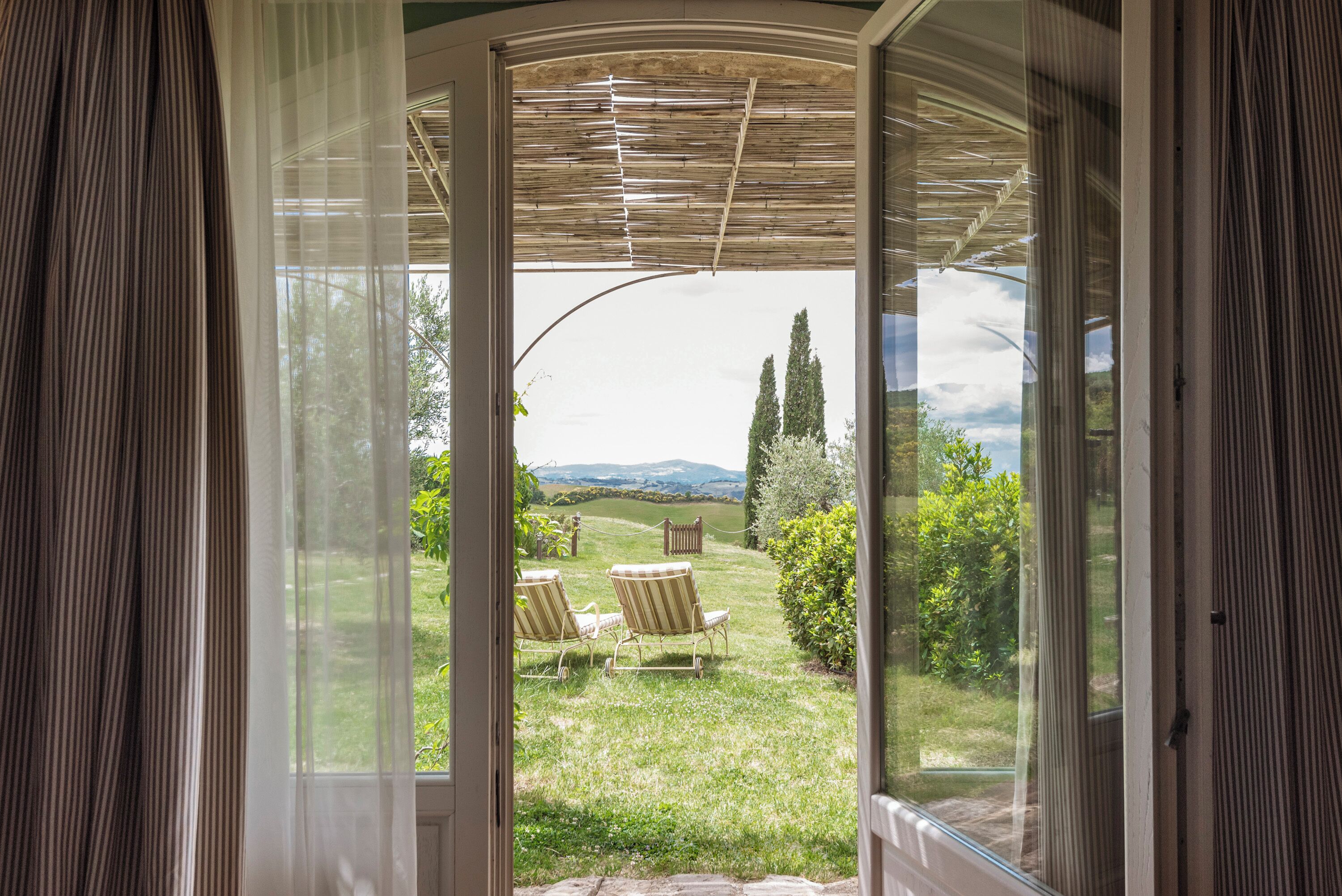 An open door to a patio with two lawn chairs and a view of a grassy landscape with trees and hills.