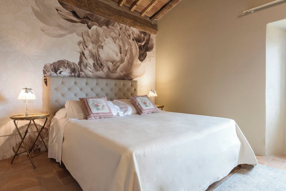Une chambre à coucher avec une tête de lit capitonnée, une grande fresque murale à motifs floraux, deux tables de chevet avec des lampes et un lit soigneusement fait avec du linge de lit blanc.