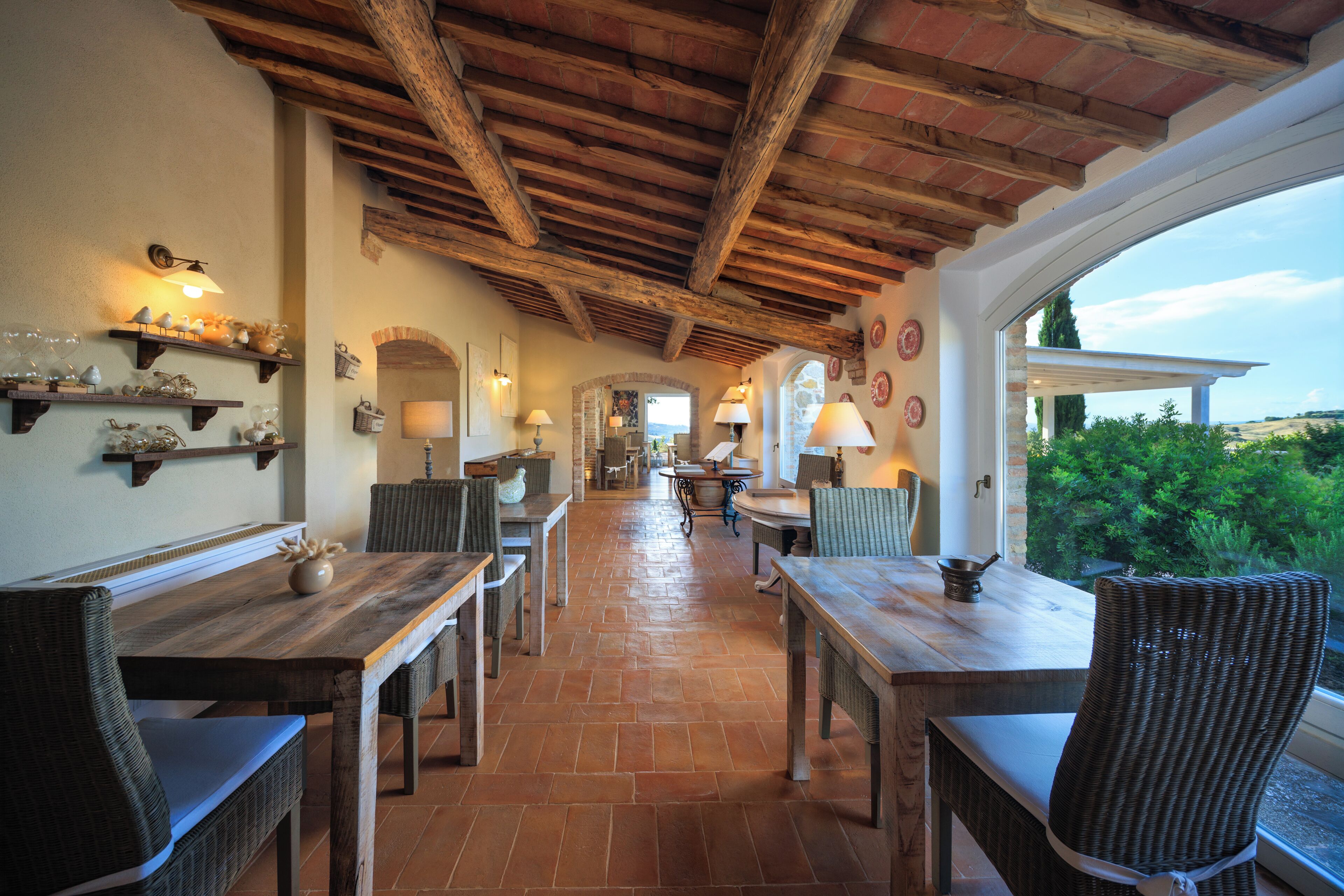 A dining area with wooden tables and wicker chairs, a brick floor, and a view through a window.