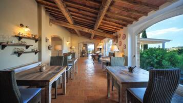 A dining area with wooden tables and wicker chairs, a brick floor, and a view through a window.