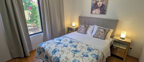 A bedroom with a bed, two bedside tables, a nightstand, a window with curtains, and a painting on the wall.