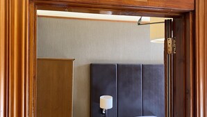 A hotel room with a wooden door labeled "TWO", a bed with white linens, a bedside lamp, and a small wooden nightstand with a lamp and a telephone.