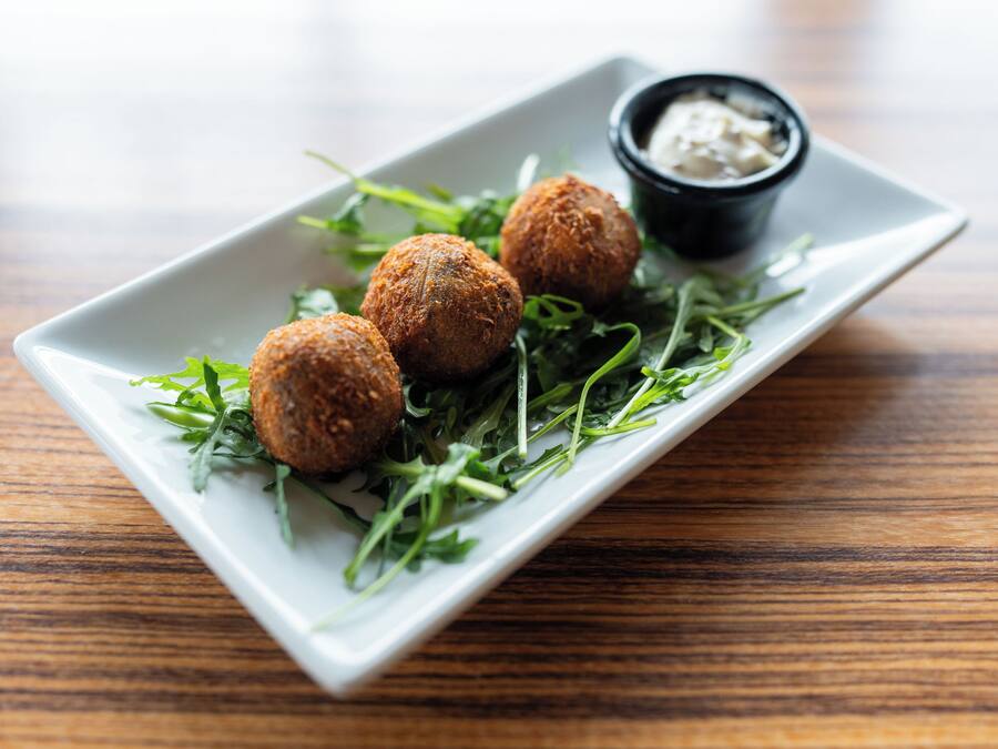 Um prato retangular branco com três bolinhas fritas redondas e douradas sobre uma base de folhas verdes, acompanhado de uma pequena tigela preta com molho branco para molhar.
