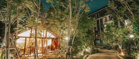 Un área de estar al aire libre con cerramiento de vidrio, rodeada de abundante vegetación y un sendero que lleva a un edificio.