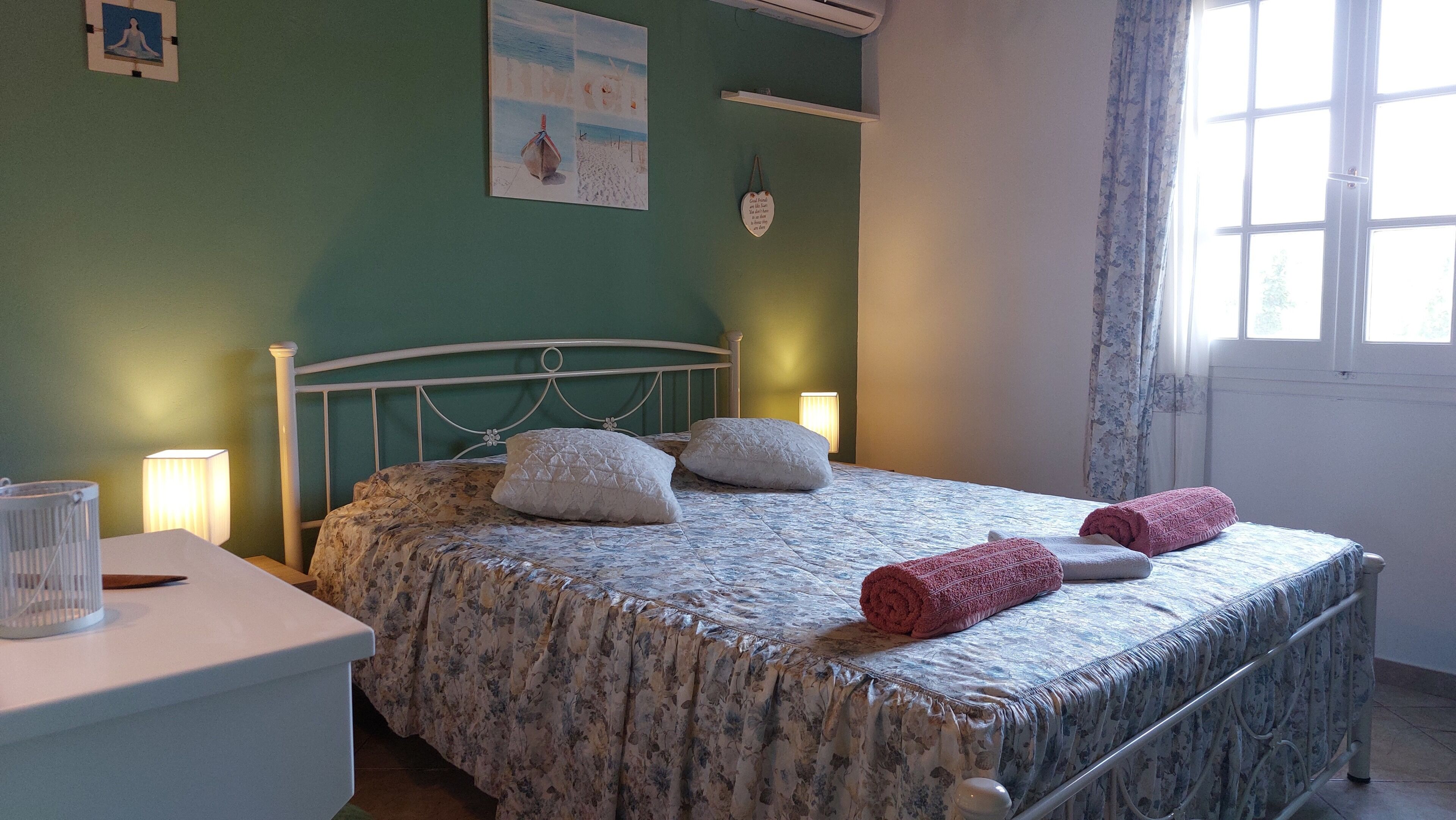 Cama bem arrumada com roupa de cama floral, mesa de cabeceira com abajur e unidade de ar condicionado na parede.