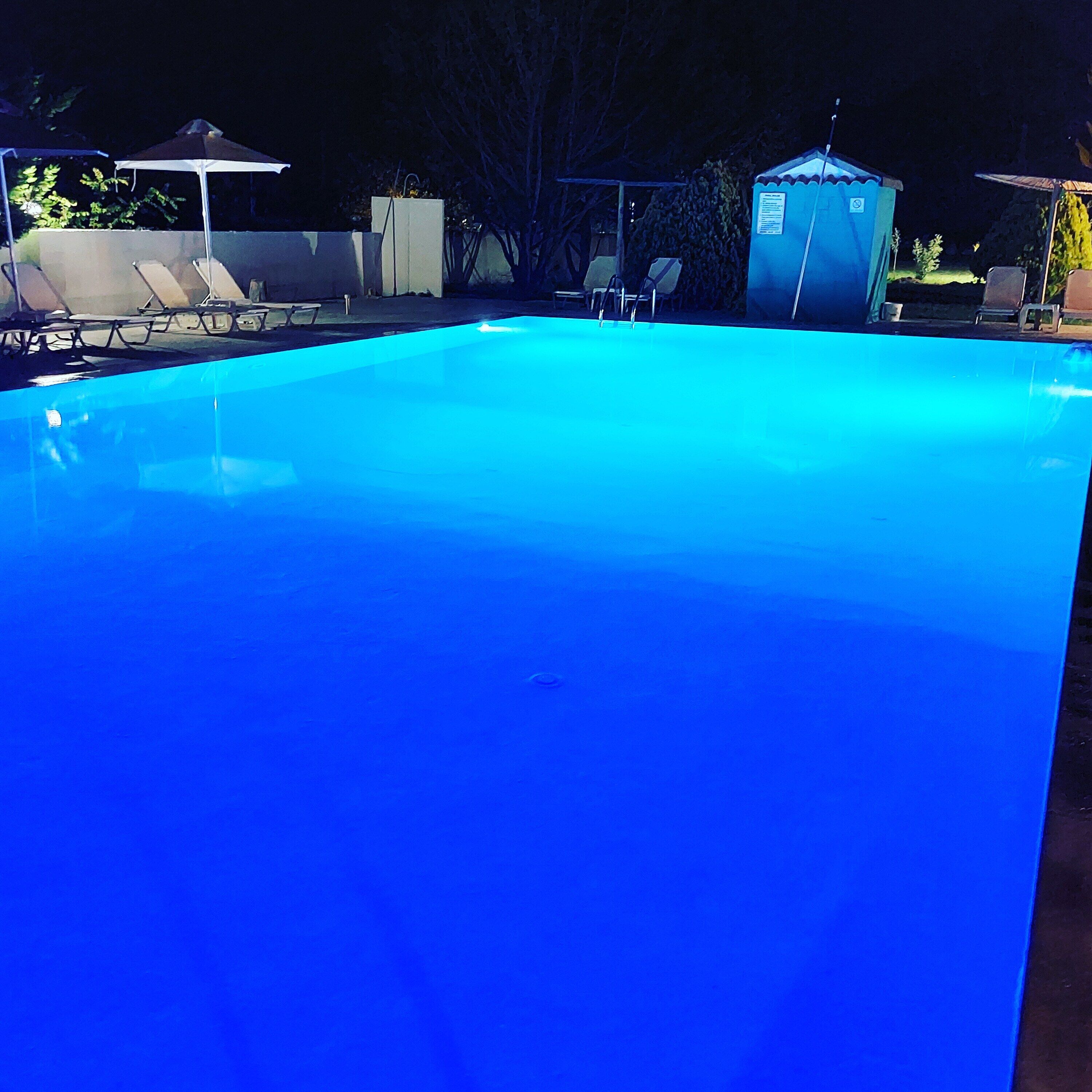 A swimming pool with a blue tint, surrounded by lounge chairs and a small shed.