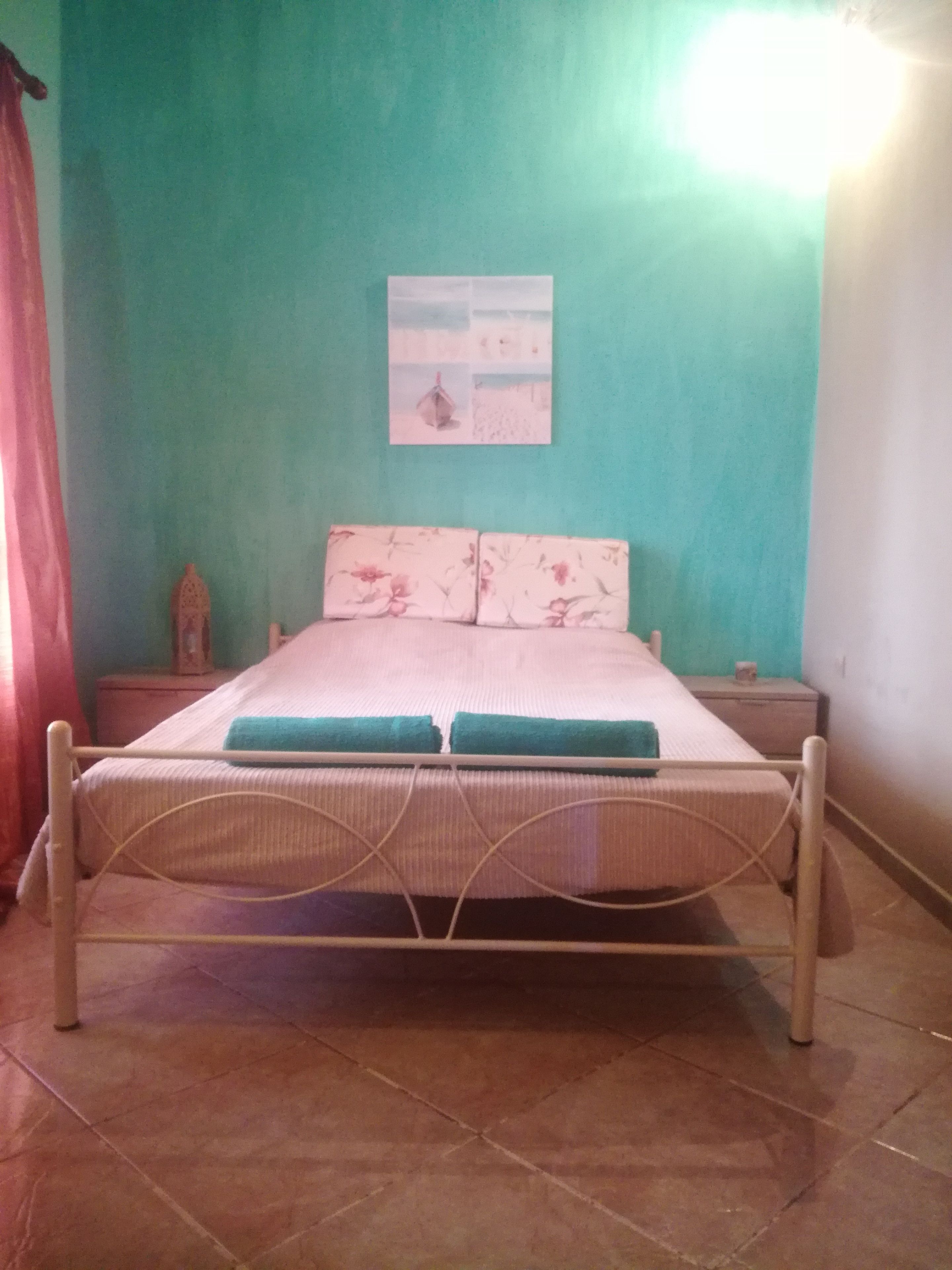 A single bed with a metal frame, a floral-patterned mattress, and two pillows. There is a small nightstand with a lamp on it to the right of the bed. The room has teal walls and a tiled floor.