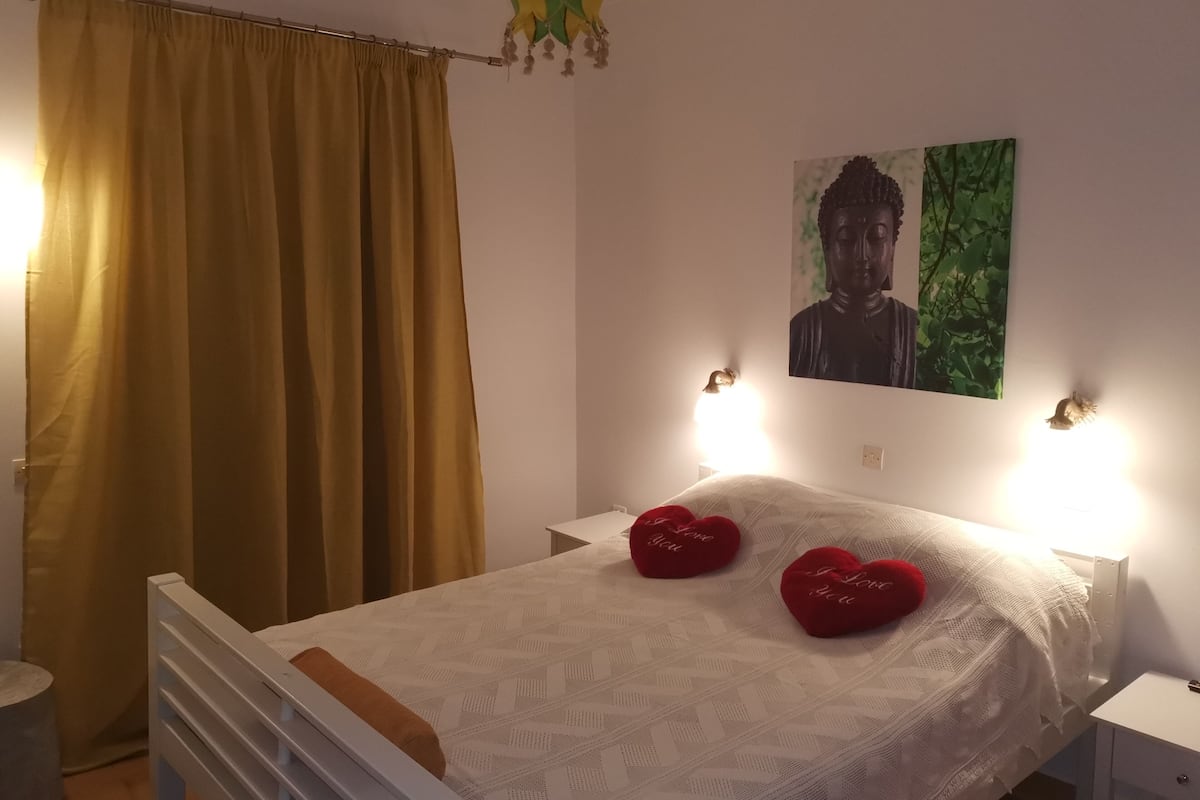 A bedroom with a bed, two bedside tables, a wall-mounted lamp, a painting, and a curtain.