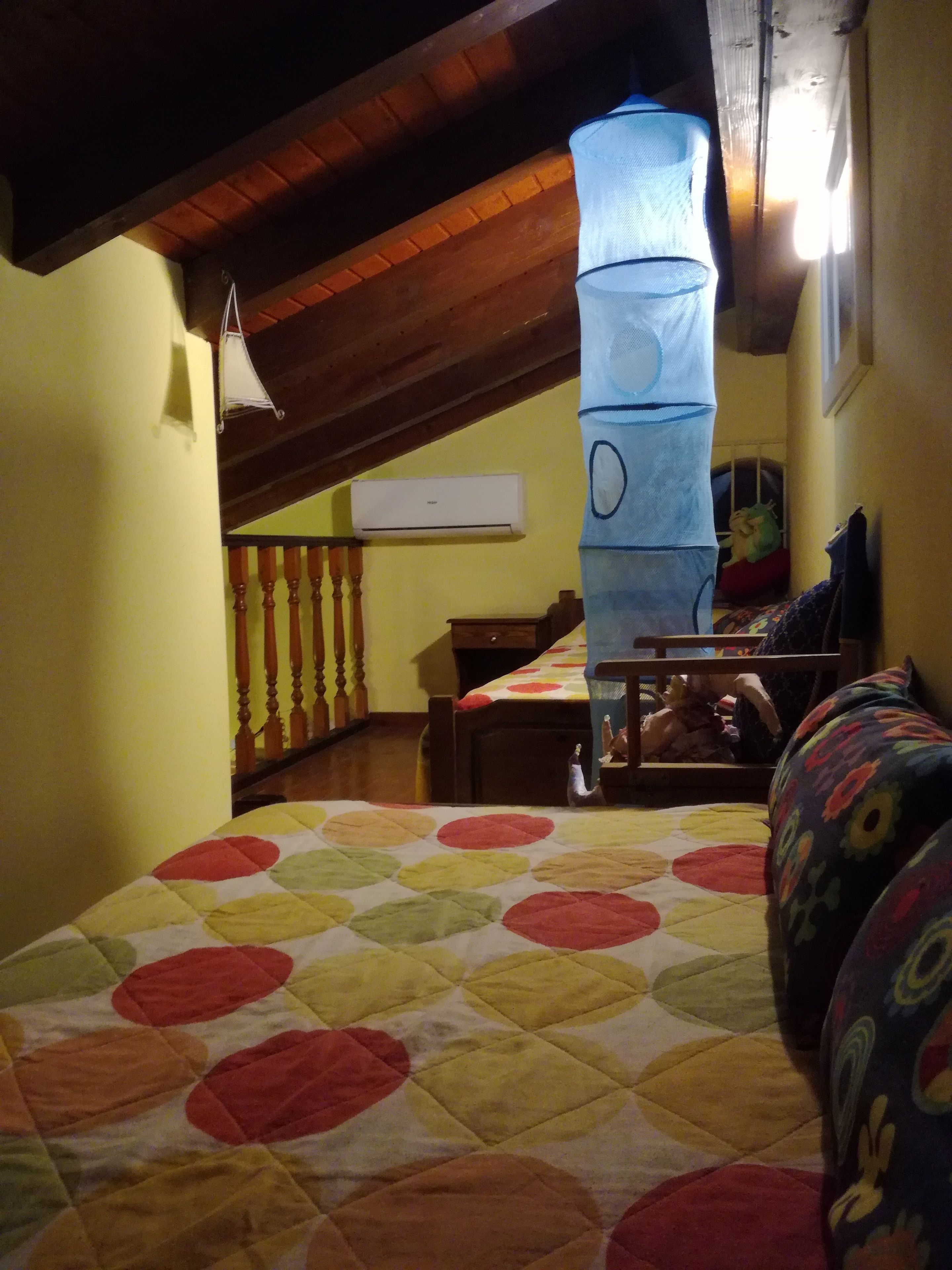 A bedroom with a bed covered in a multicolored quilt, a wooden ceiling, a window with a view, and a wall-mounted air conditioner.