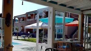 An outdoor dining area with a table and chairs, a glass door leading to a pool, and umbrellas. - Sausan Hotel (Corfu)