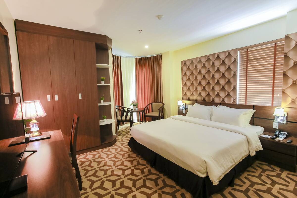 A hotel room with a large bed, a desk with a lamp, a chair, a small table with a plant, and a wardrobe.