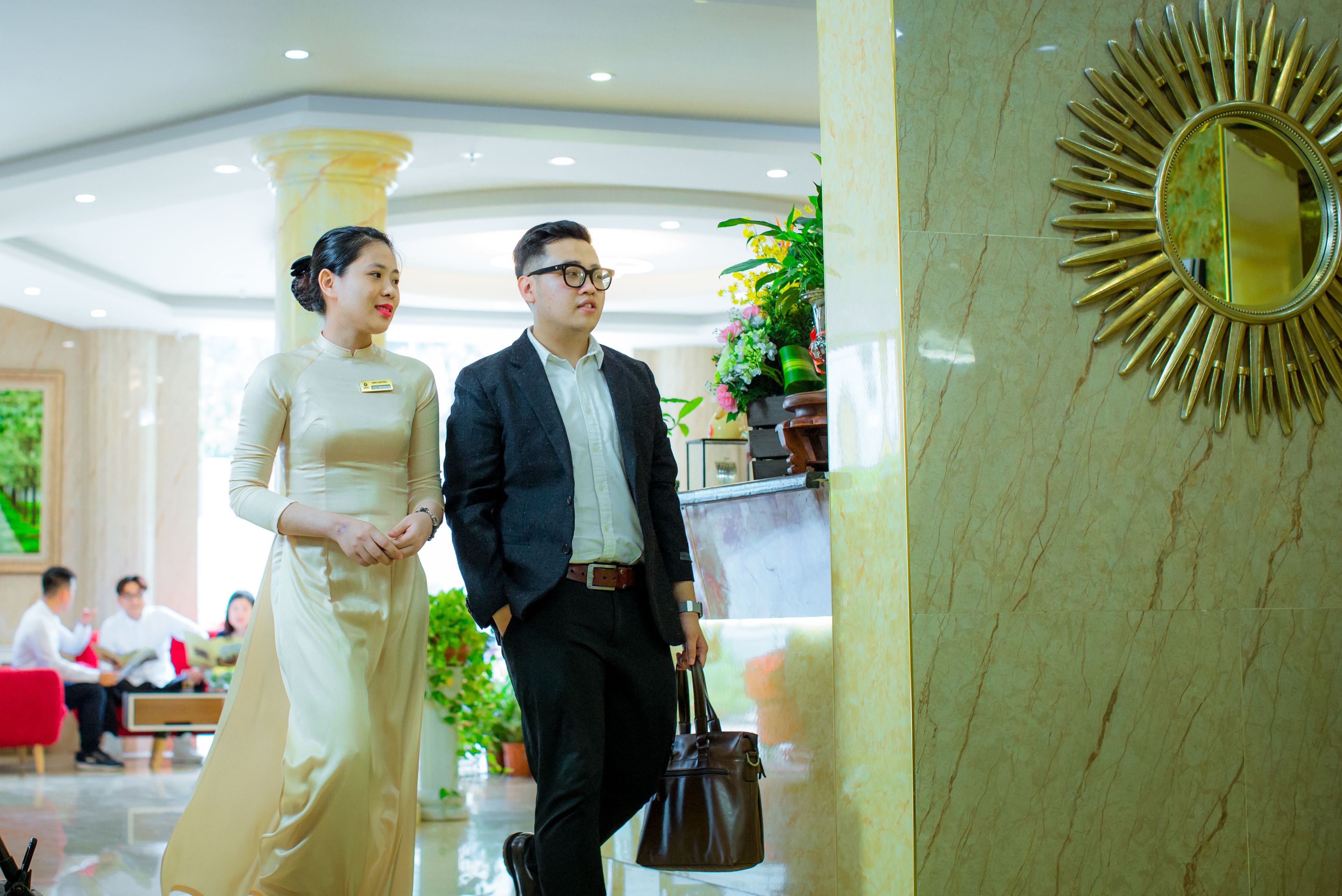 A man and a woman walking in a hotel lobby with marble floors and a sunburst mirror.