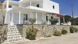 A two-story white building with a stone wall in front, a covered porch, and a small garden with potted plants. - Kirki Studios (Paros)