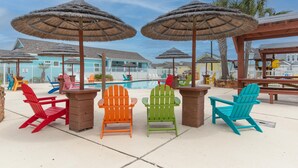 Uma área à beira da piscina com cadeiras Adirondack coloridas e guarda-sóis de palha, uma piscina e casas de praia ao fundo.