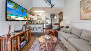 Uma sala de estar moderna com uma televisão de tela plana grande, uma mesa de centro de madeira, um sofá e uma cozinha ao fundo.
