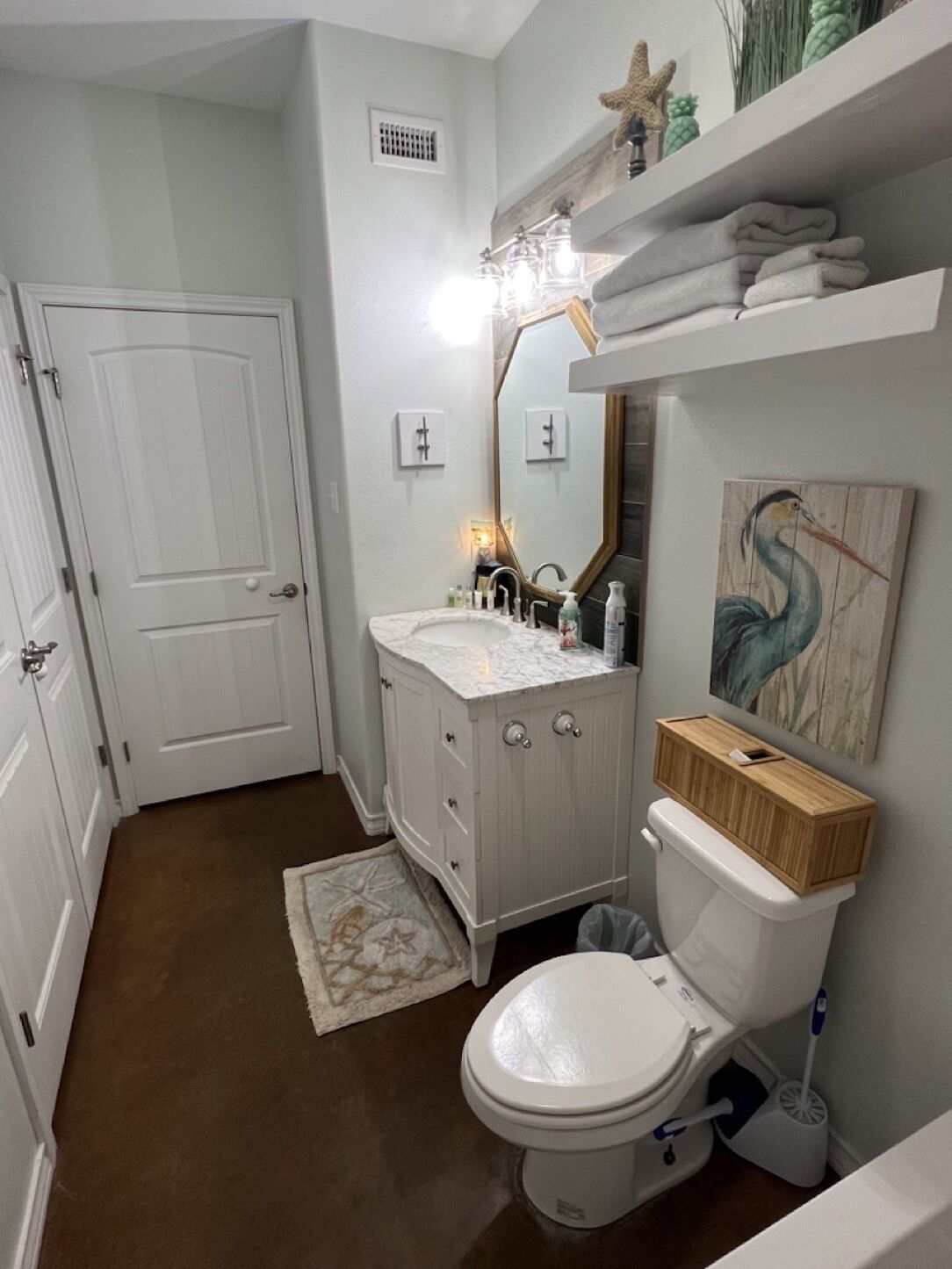 A bathroom with a white toilet, a vanity with a mirror, and a wooden shelf above the toilet.