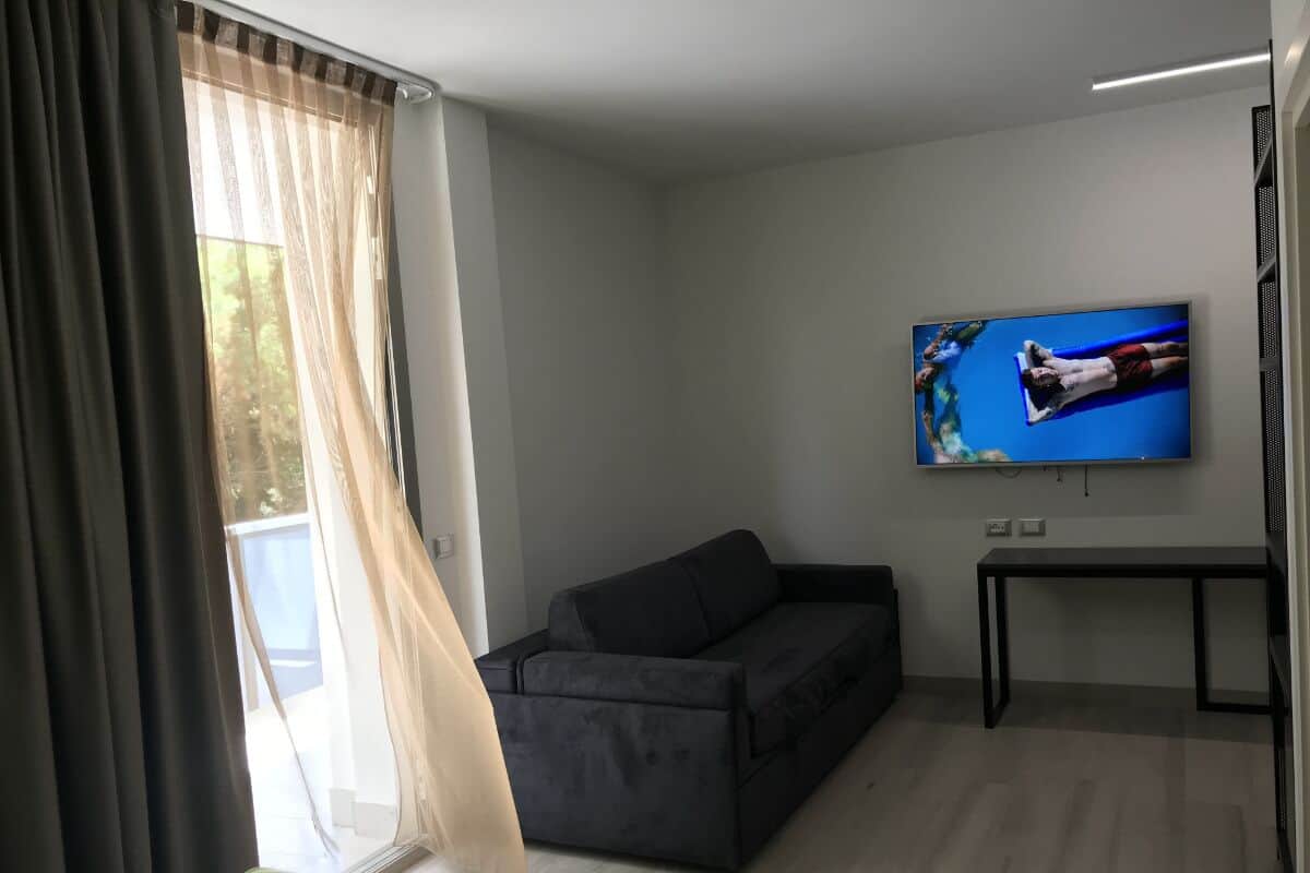 Una sala de estar moderna con un sofá negro, un televisor montado en la pared y una ventana con cortinas de gasa.
