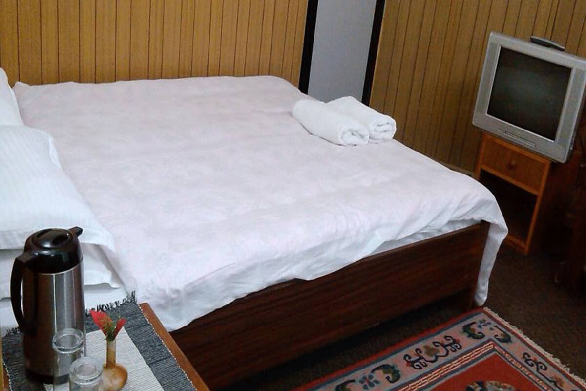 A neatly made bed with a white sheet and a rolled towel, a wooden bedside table with a television, and a small table with a coffee pot and glasses.