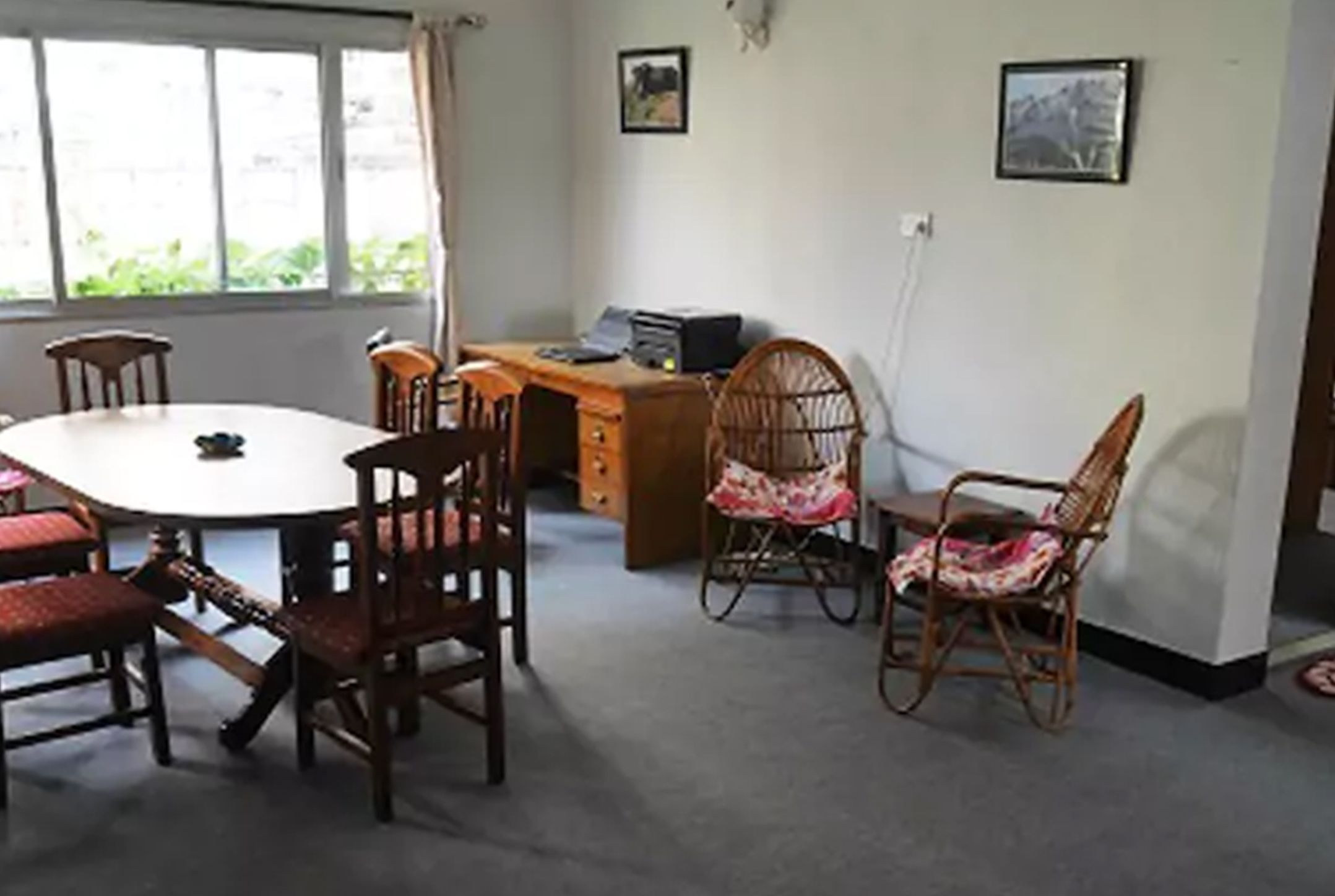 A room with a round table, four wooden chairs, a desk with a computer, and two framed pictures on the wall.