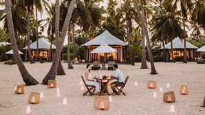 Un área de comedor frente a la playa con mesas y sillas, rodeada de palmeras y cabañas de playa.