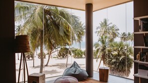 Una sala de estar moderna con un amplio ventanal que da a una playa, con suelo de madera, sillones tipo flexo y una estantería.