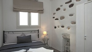 A bedroom with a bed, a bedside table with a lamp, a window with curtains, and a wall with a rock design. - Portes View House (Paros)