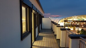 A wooden deck with a railing, a glass door, and a view of a building with a curved roof and signage. - RheinCity Hotel (Ludwigshafen)