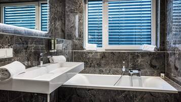 A modern bathroom with a large mirror, two sinks, a bathtub, and a window with blinds.
