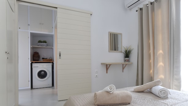 A bedroom with a bed, a washing machine, a mirror, and a shelf with a plant. - Vagia Calm House (Paros)