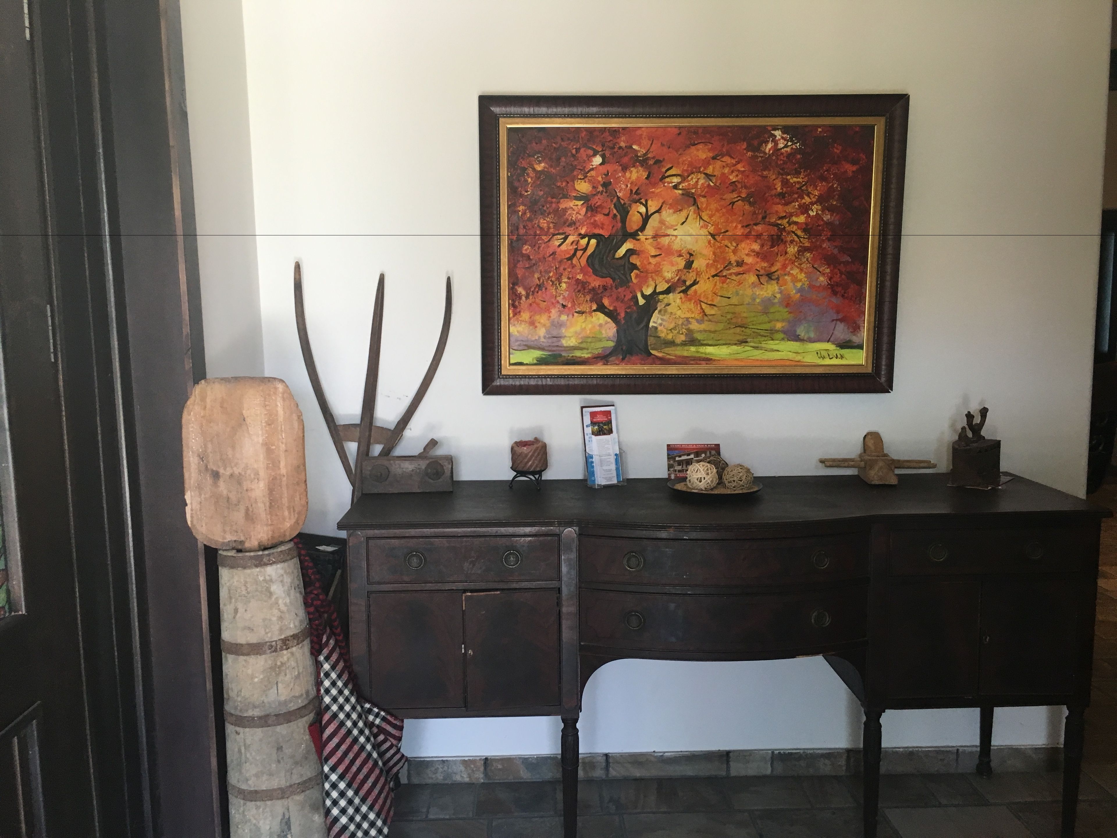 Uma mesa de madeira com gavetas, um quadro emoldurado de uma árvore e uma pequena prateleira com objetos decorativos.