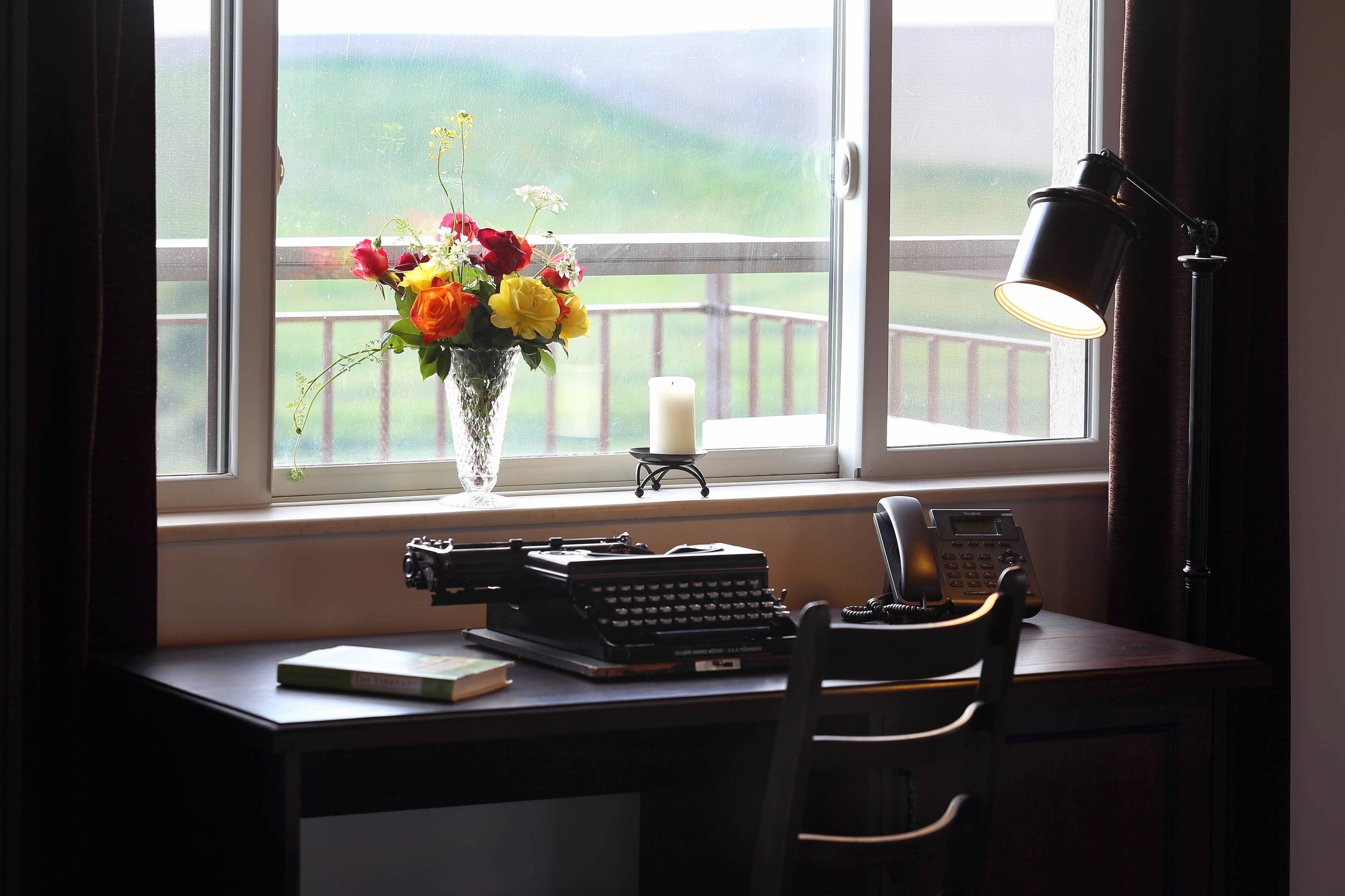 Un bureau avec une machine à écrire, un téléphone et un vase de fleurs, placé près d’une fenêtre donnant sur une terrasse.