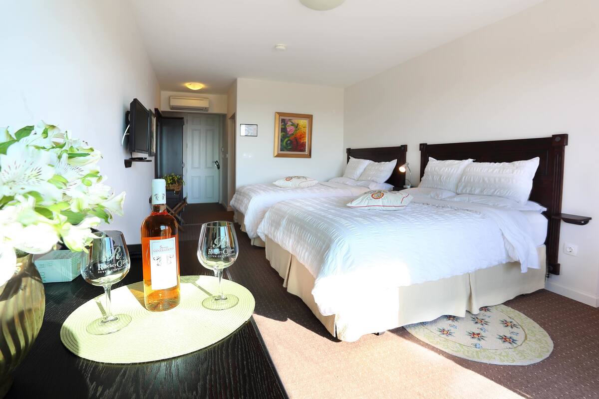 Une chambre d’hôtel avec deux lits, un téléviseur fixé au mur, un tableau et une table sur laquelle se trouvent une bouteille de vin et un verre.