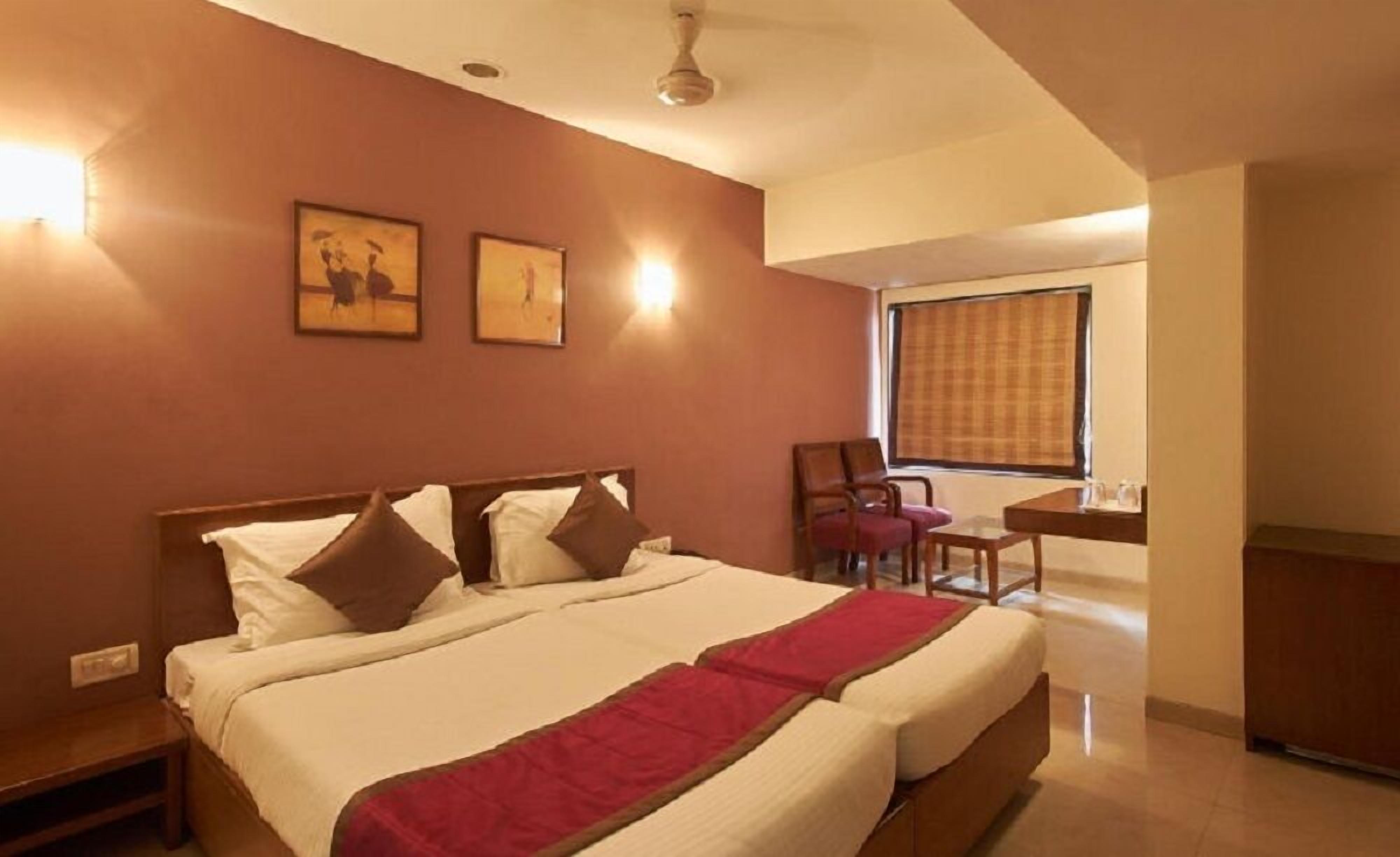 A hotel room with a large bed, a desk, and a chair. There are two wall-mounted lights and a window with blinds.