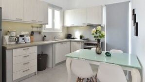 A modern kitchen with light-colored cabinets, a glass-top dining table, white chairs, a trash bin, and a vase with flowers. - The Local House (Paros)