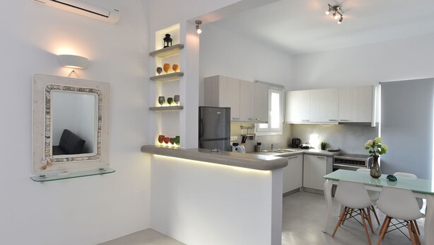 A modern kitchen with white cabinets, a built-in refrigerator, and a dining area with a glass table and chairs. - The Local House (Paros)