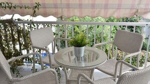 A round glass table with wicker chairs and a potted plant on a balcony with a white railing and greenery in the background. - The Local House (Paros)