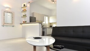 A modern living room with a black sofa, a white coffee table, and a small round table. There is a kitchen in the background with white cabinets and a black countertop. - The Local House (Paros)