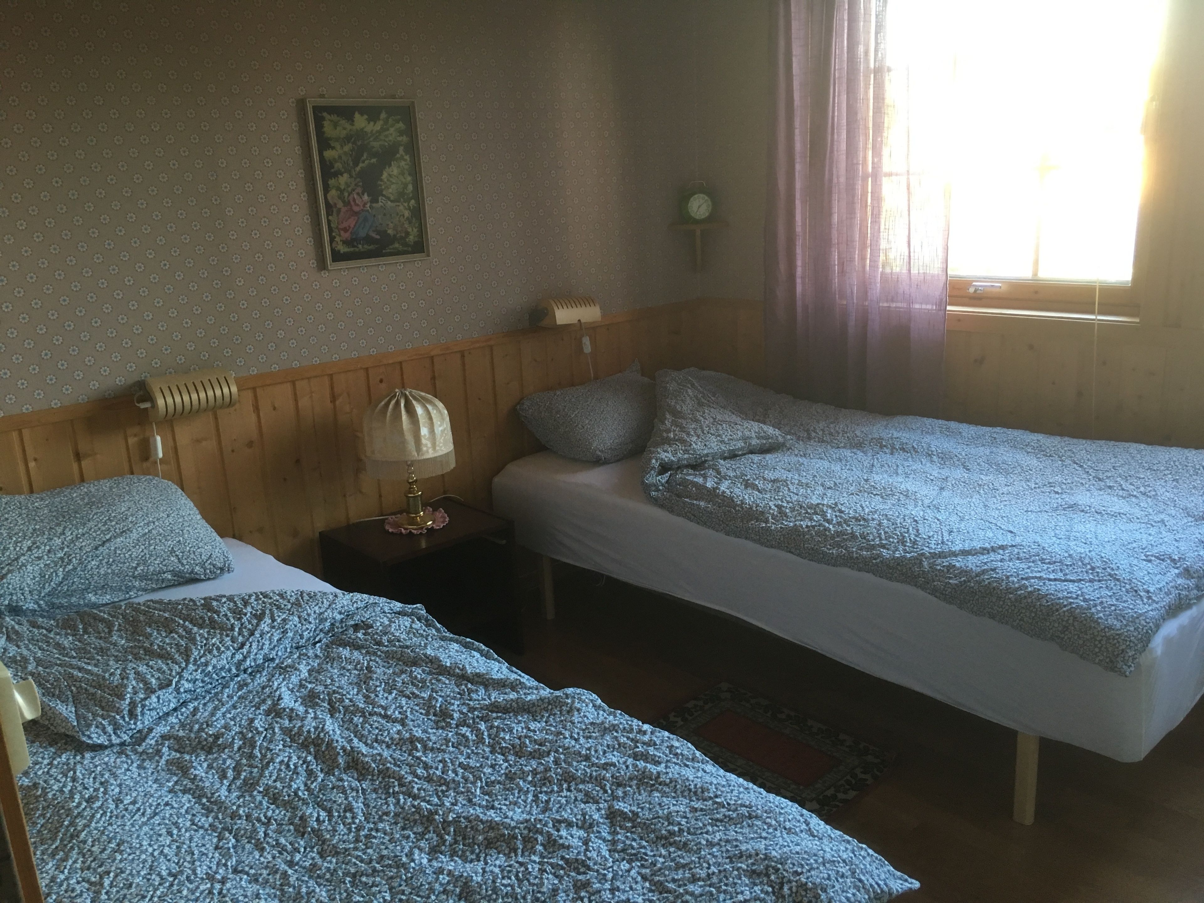 A room with two single beds, a wooden headboard, a small bedside table with a lamp, a framed picture on the wall, and a window with curtains.