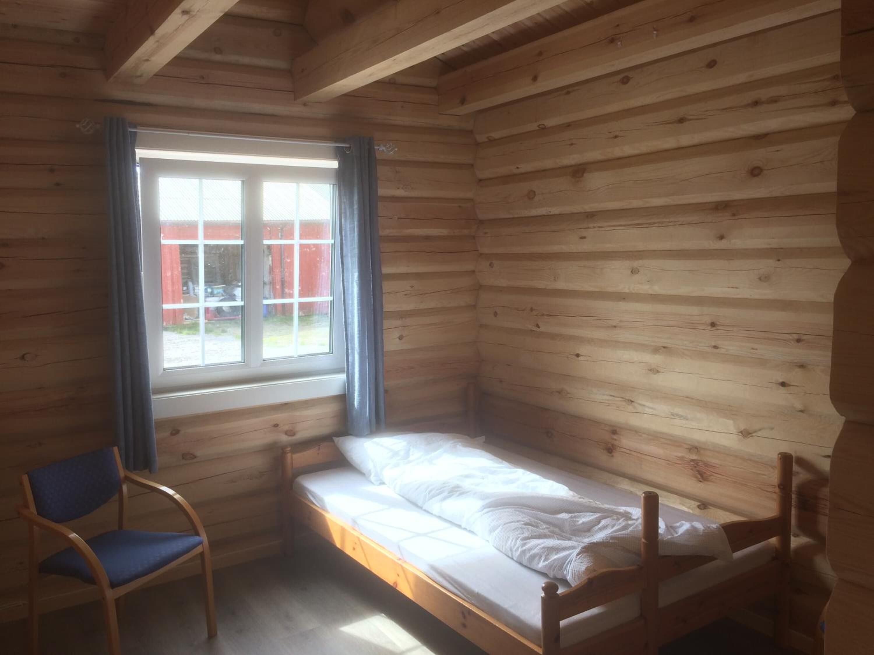 Ein kleines Zimmer mit Holzwand, einem Einzelbett, einem Stuhl und einem Fenster mit Vorhängen.