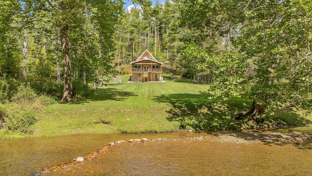 A two-story wooden cabin with a balcony, surrounded by a forested area and a small stream. - Retriever Beach - New River-frontage with Your Own Private Beach! Fire Pits - Games and More! (West Jefferson)