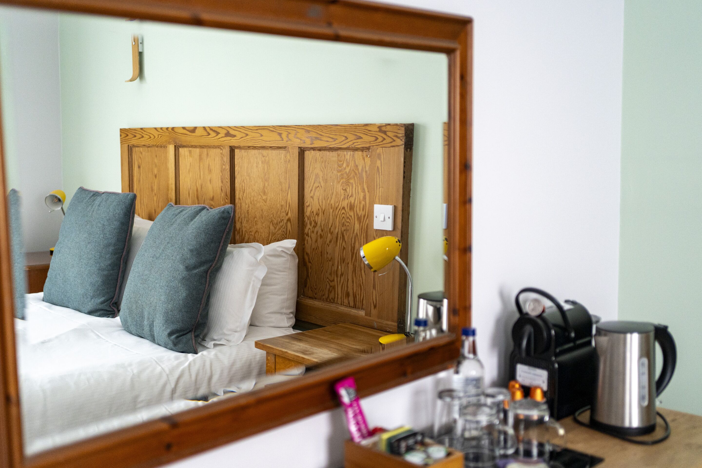 Une chambre d’hôtel avec une tête de lit en bois, un lit avec une literie grise et blanche, une table de chevet avec une lampe, une machine à café et un miroir qui reflète la pièce.